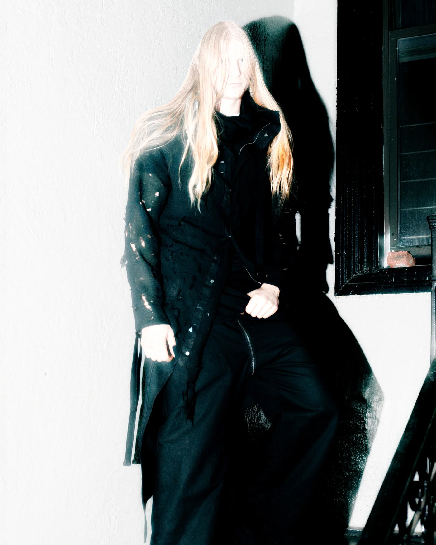 The designer of Synthesis Documentation wearing a handmade black coat, photographed against a white wall.