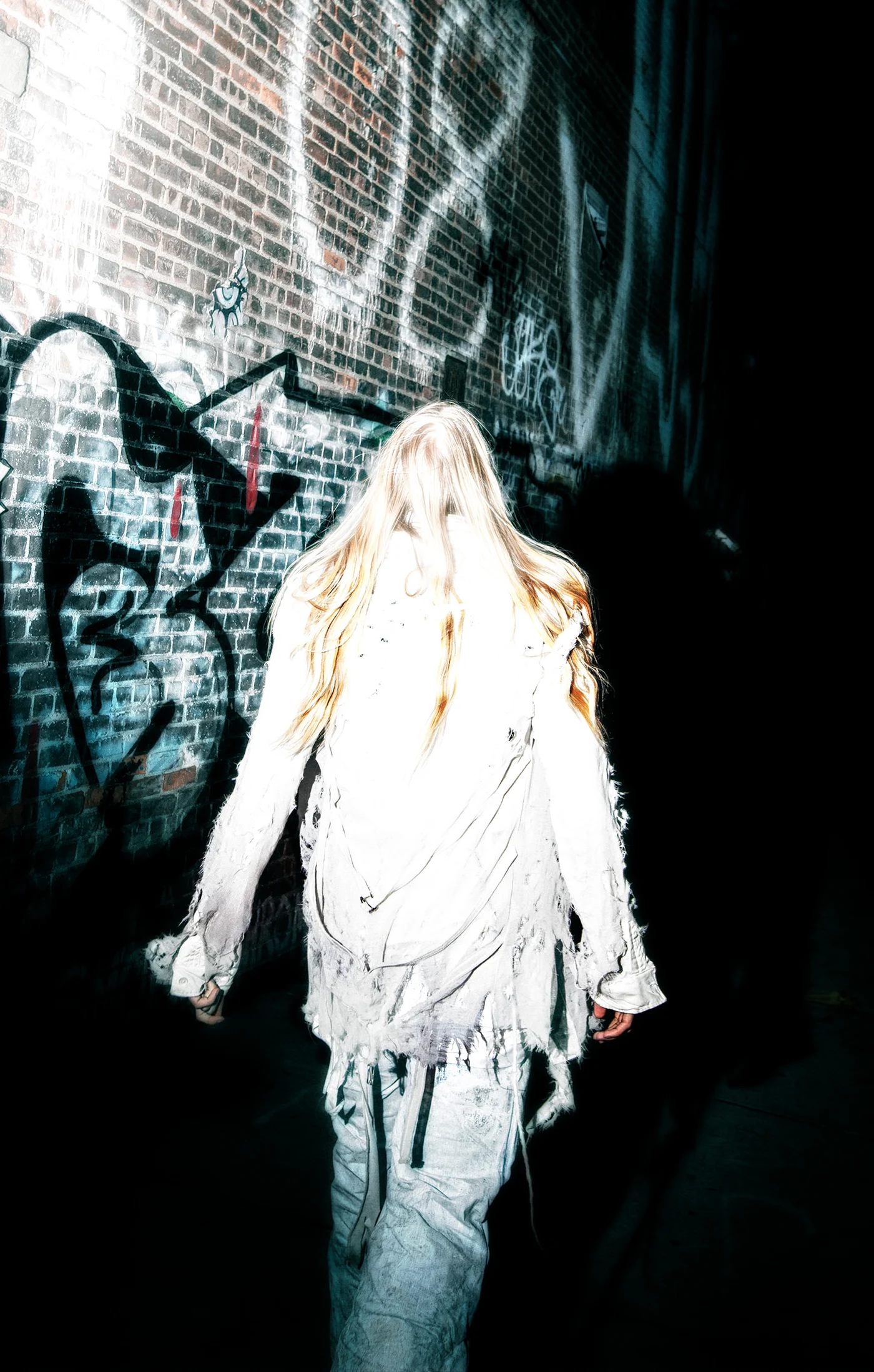 The Synthesis designer wearing a white deconstructed garment beside a brick wall.